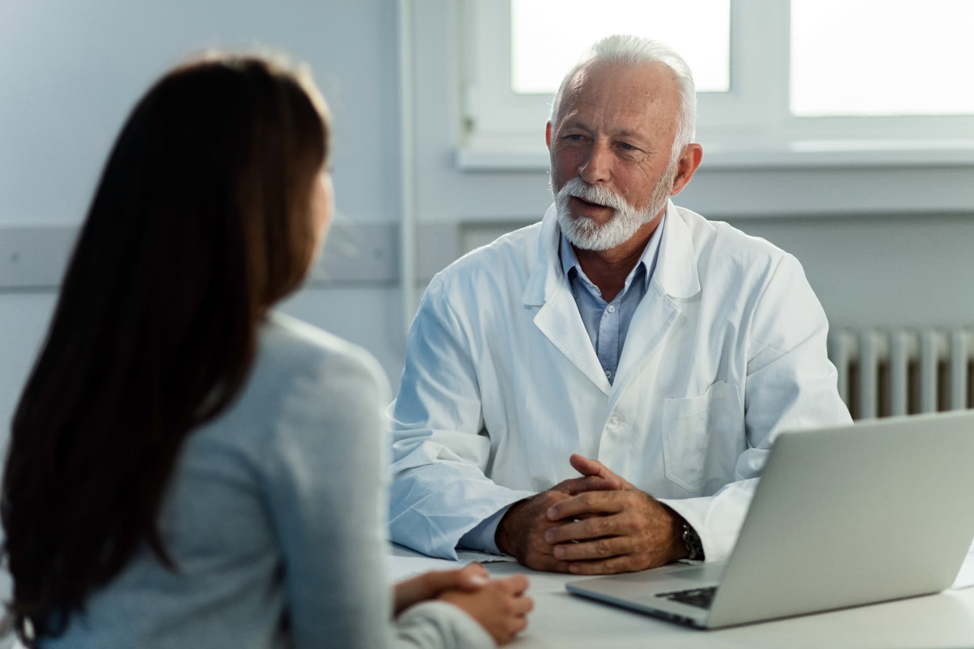 Médecin sénior en blouse blanche échangeant avec une patiente en cabinet médical, ordinateur portable posé sur le bureau.